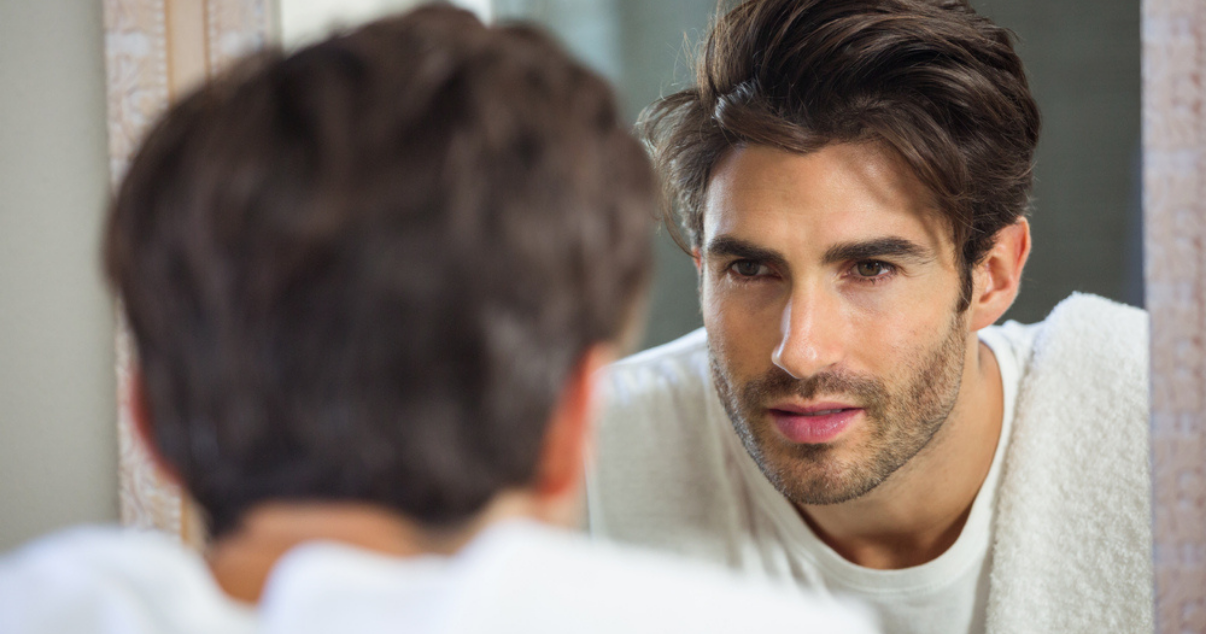 Young man looking into the mirror, deciding to repair his Hair System or not